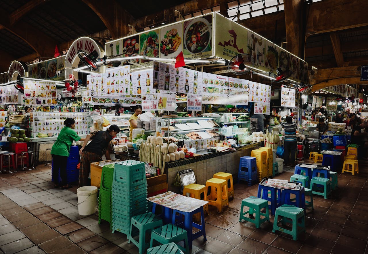 About A vibrant food stall in a busy Vietnamese market with colorful seating and various food displays.